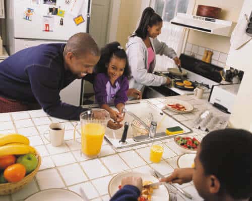 Black family prepares for breakfast together in the kitchen. Mother makes pancakes at stove, while father helps child wash hands as other child looks on.